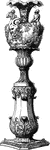 This flower stand with vase is a maltese design. The stand has a simple leaf design with a base that rests on animal claws, while its top flares out. The vase is richly designed with flowers and leaves and unusual human figures seated on each side, as well as, a large horned human head.